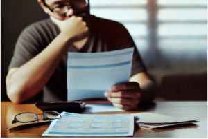 Imagem de uma pessoa sentada à mesa, analisando documentos financeiros com expressão pensativa e preocupada. O ambiente é simples, com papéis, contas e uma calculadora sobre a mesa, representando a realidade de alguém lidando com dívidas e buscando soluções. A imagem transmite o conceito da Lei do Superendividamento no Brasil, que oferece suporte para pessoas em situação de endividamento, destacando a importância do alívio e da orientação financeira.