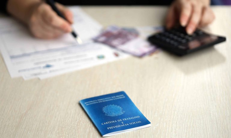 Homem fazendo contas em cima de uma mesa onde possui uma carteira de trabalho