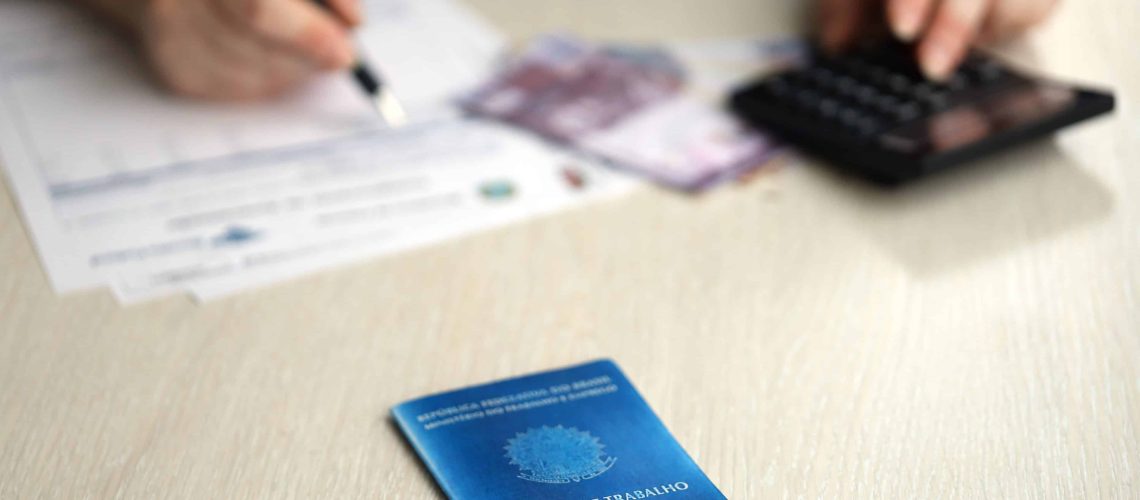 Homem fazendo contas em cima de uma mesa onde possui uma carteira de trabalho